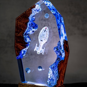 May include: An illuminated resin sculpture with a white spaceship model encased in clear resin. The sculpture has blue and brown accents, and a wooden base. The spaceship is a key feature of the design.