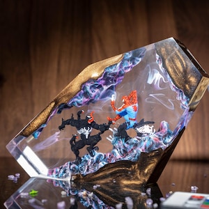 May include: A clear resin lamp featuring a battle scene with Spider-Man and Venom figures. The lamp has a gold-colored border and a swirling purple and blue design within the resin. A remote control is visible.