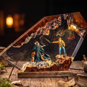 May include: A trapezoidal resin and wood lamp featuring a miniature scene of two figures dueling with wands. One figure wears a dark green robe with a snake, while the other wears a yellow jacket and blue jeans. Wisps of smoke swirl inside the clear resin.