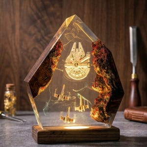May include: A pentagonal resin lamp featuring a wooden base and embedded miniature starships, including a Millennium Falcon and X-wing fighters, suspended within clear resin flanked by textured, dark, reddish-brown wood edges.