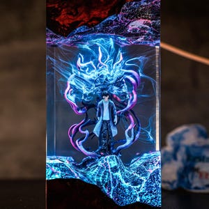 May include: A rectangular resin sculpture featuring a figure in a long coat, surrounded by blue and purple energy effects. The sculpture has a dark base and a red and black top, creating a dramatic, illuminated display.
