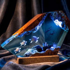 May include: A decorative lamp featuring a slanted block of wood and clear resin. Inside the resin are three sea turtles, two scuba divers, a small shipwreck, and blue LED lighting that illuminates the underwater scene from within the wood.