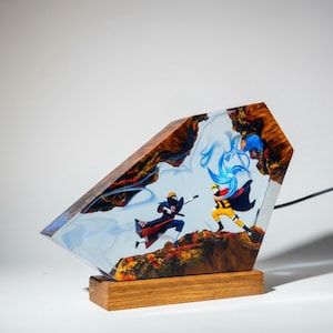 Pu&ograve; includere: Una lampada decorativa composta da un blocco di legno levigato e resina trasparente con figure anime incorporate. La scena raffigura due personaggi in una posa di combattimento dinamica, circondati da effetti di fumo in resina blu.