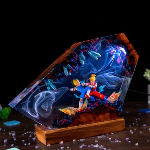 Pu&ograve; includere: Una lampada decorativa con base in legno e un blocco di resina contenente due personaggi anime in una posa di combattimento dinamica. La resina presenta effetti di fumo bianco vorticoso, accenti blu e rossi e forme cristalline illuminate.