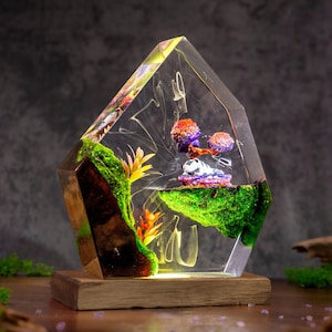 May include: A clear, angular resin sculpture containing a miniature landscape with green moss, orange and yellow plastic plants, and a small white and purple creature resting on a floating island, all mounted on a rectangular wooden base.