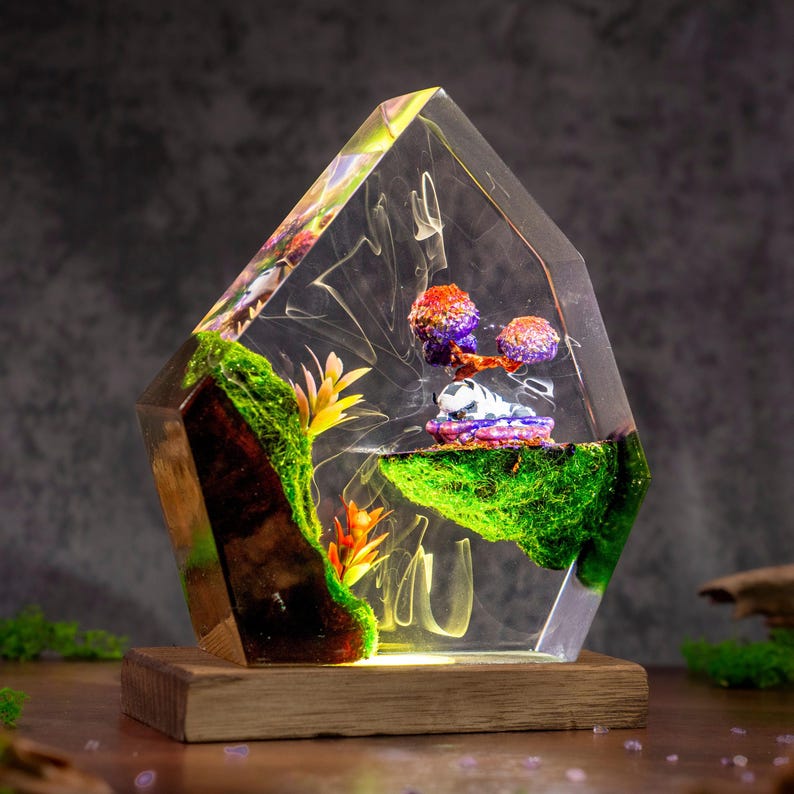 May include: A clear, angular resin sculpture containing a miniature landscape with green moss, orange and yellow plastic plants, and a small white and purple creature resting on a floating island, all mounted on a rectangular wooden base.