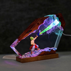 May include: A decorative lamp featuring a miniature figure with spiky green hair and white pants, positioned on a wooden base. The figure is encased in a dark wood frame with translucent resin, emitting a glowing green and purple light effect.
