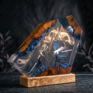 May include: A rectangular resin and wood lamp featuring two miniature figures, Cloud and Sephiroth, in a combat pose. The clear resin contains swirling white smoke effects, while the edges are framed by natural wood and blue crystal-like accents.