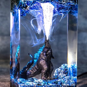 May include: A clear resin block with a blue and white swirling pattern. A black Godzilla figurine is inside the block, with its mouth open and smoke rising from it.