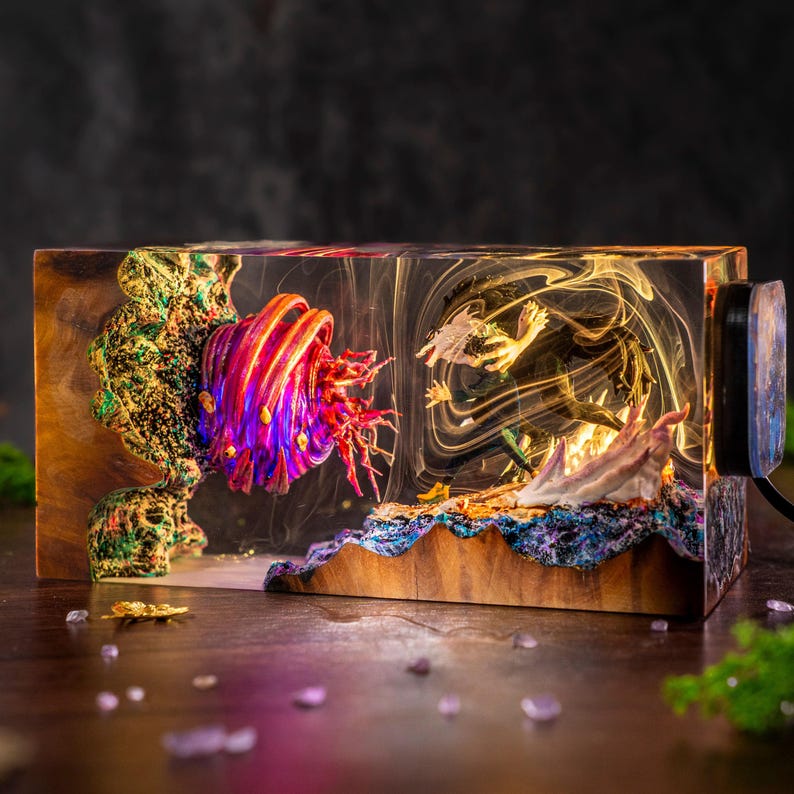May include: A rectangular resin and wood lamp featuring a vibrant, multi-colored spherical creature with tentacles on the left and a dynamic, dark-haired figure in a combat pose on the right, all encased in clear resin with swirling white smoke effects.