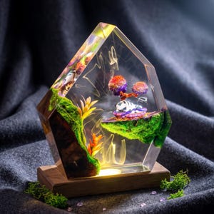 May include: A pentagonal resin sculpture featuring a miniature landscape with green moss, orange and yellow plastic plants, and a small white and black creature resting on a floating island, all set on a rectangular wooden base with internal lighting.