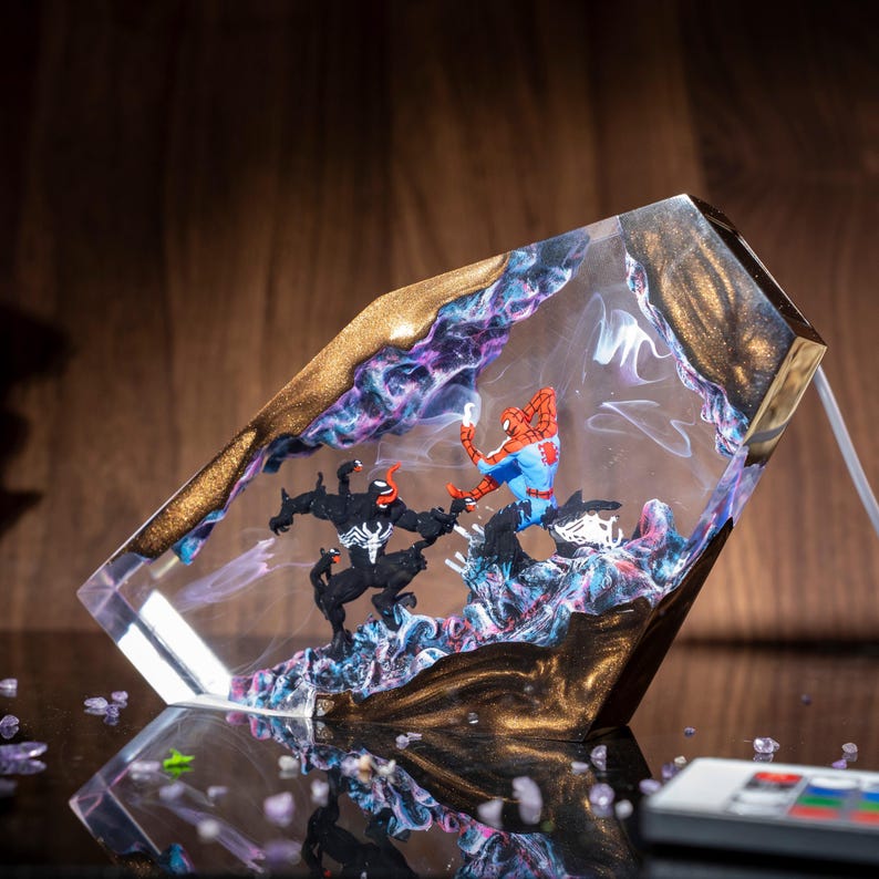 May include: A clear resin lamp featuring a battle scene with Spider-Man and Venom figures. The lamp has a gold-colored border and a swirling purple and blue design within the resin. A remote control is visible.