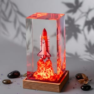 May include: A rectangular clear resin block containing a miniature space shuttle model with a bright orange and red illuminated exhaust plume at the base, mounted on a square wooden stand with a smooth, light-colored grain finish.