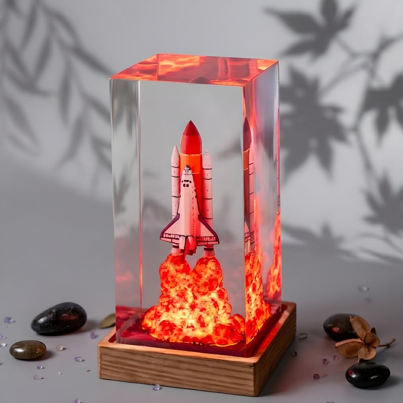 May include: A rectangular clear resin block containing a miniature space shuttle model with a bright orange and red illuminated exhaust plume at the base, mounted on a square wooden stand with a smooth, light-colored grain finish.