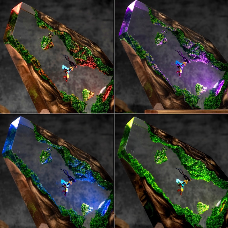May include: A clear resin sculpture shaped like a crystal, featuring embedded dark wood, vibrant green moss-like textures, and a small figure in a blue and yellow outfit. The piece is illuminated with changing red, purple, blue, and green LED lights.