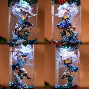 May include: A rectangular resin block containing a miniature figure of a blonde character in blue tunic and cape, holding a shield above their head. White swirling resin effects surround the figure, which stands atop a green and grey base.