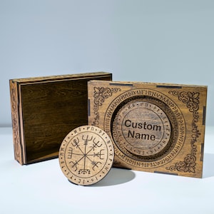 May include: A rectangular wooden box with an open lid and a circular wooden coaster. The box lid features a circular engraving with the text Custom Name surrounded by runes. The coaster displays a Vegvisir symbol surrounded by a runic border.