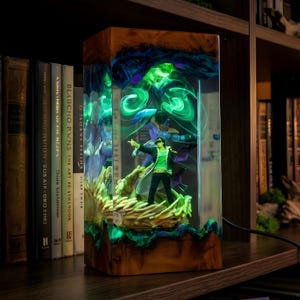 May include: A rectangular wooden lamp with a clear resin front, showcasing a figurine illuminated by green and blue lights. The lamp features a detailed figure and swirling patterns, creating a unique decorative piece.