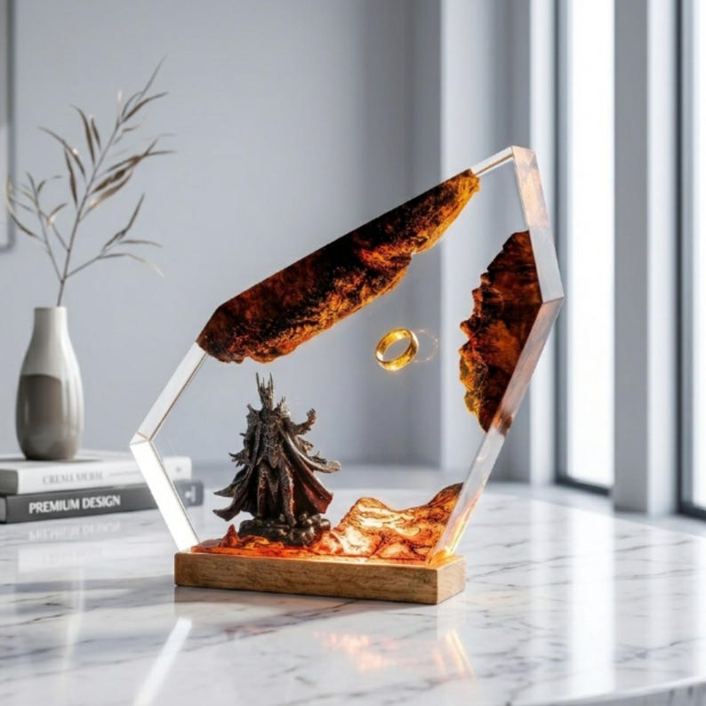 May include: A decorative display piece featuring a dark armored figure standing on a textured orange base, enclosed within a clear geometric resin frame with embedded wood-like elements and a single gold ring suspended in the center.