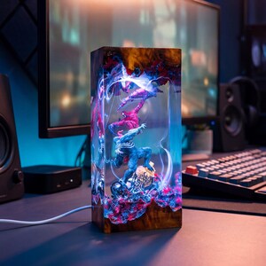 May include: A rectangular lamp featuring superhero figures encased in clear resin. The lamp has a wooden base and displays a vibrant scene with red, blue, and purple hues. The figures are illuminated by internal lighting, creating a dynamic display.
