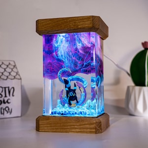 May include: A rectangular resin lamp featuring a black Umbreon figure encased in clear resin with swirling purple and blue smoke effects. The lamp is framed by top and bottom wooden blocks and illuminated from within, creating a glowing display.
