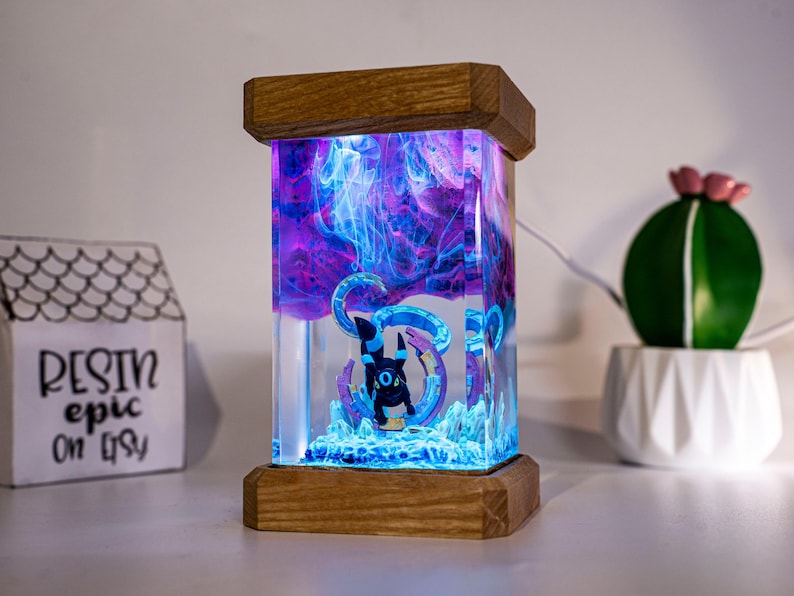 May include: A rectangular resin lamp featuring a black Umbreon figure encased in clear resin with swirling purple and blue smoke effects. The lamp is framed by top and bottom wooden blocks and illuminated from within, creating a glowing display.