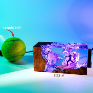 May include: A tennis ball next to a wooden block with a colorful, illuminated scene inside. The scene depicts a figure in a dark suit. The block is labeled "SIZE M".