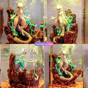 May include: A clear resin block containing a white and purple Mewtwo figure standing on dark red rocky terrain with bright green lightning-like accents. The interior features swirling yellow smoke effects and the text ResinEpic on Esty.