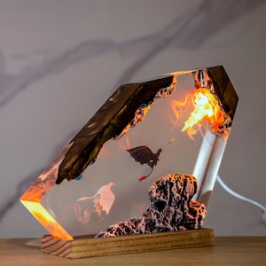May include: A rectangular resin lamp featuring a dark, jagged rock-like top and a textured base. Inside the clear resin, a black dragon and a white dragon are suspended in flight near a glowing orange light source resembling fire and smoke.