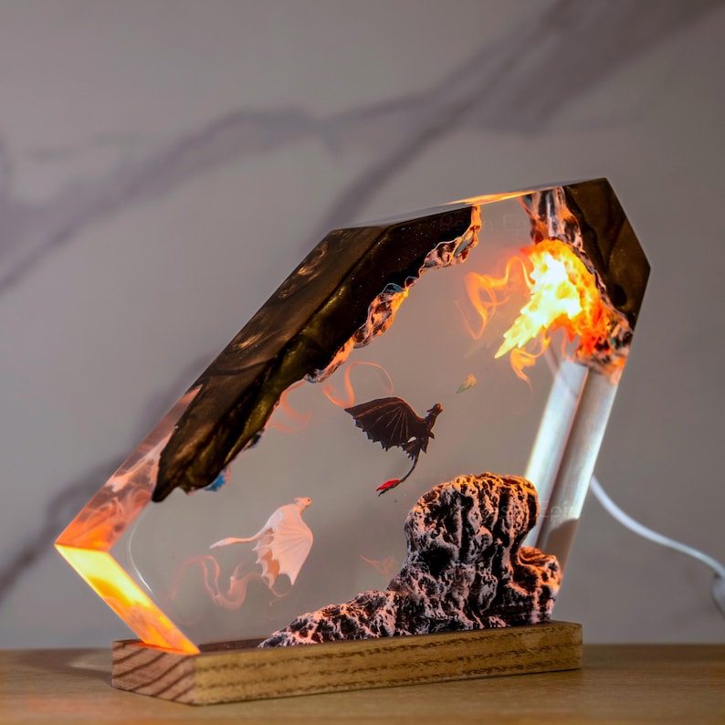 May include: A rectangular resin lamp featuring a dark, jagged rock-like top and a textured base. Inside the clear resin, a black dragon and a white dragon are suspended in flight near a glowing orange light source resembling fire and smoke.