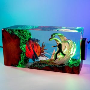 May include: A rectangular resin and wood lamp featuring a miniature figure in a black suit standing amidst swirling white energy effects. To the left, a dark wood section glows with green light, while a red and blue textured orb sits nearby.