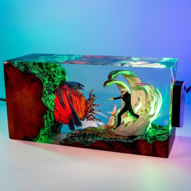 May include: A rectangular resin and wood lamp featuring a miniature figure in a black suit standing amidst swirling white energy effects. To the left, a dark wood section glows with green light, while a red and blue textured orb sits nearby.