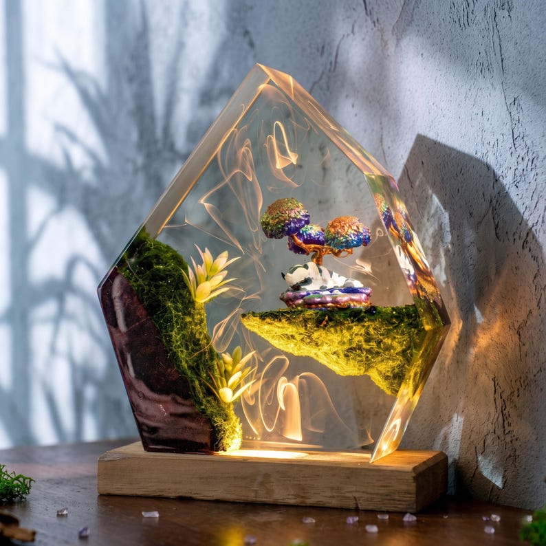 May include: A pentagonal resin lamp featuring a clear, angular frame with internal swirling patterns. Inside, a miniature iridescent tree and a small white animal figure rest on a mossy ledge, illuminated from a rectangular wooden base.