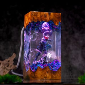 May include: A rectangular wooden lamp with a resin core featuring a Venom figurine. The wood has a natural grain, and the resin is filled with swirling blue and purple colors. The figurine has red accents and is posed on a rocky base.