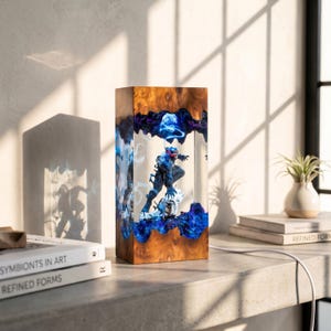 May include: A rectangular wooden lamp with a superhero figure encased in clear resin. The lamp features a wood grain exterior, blue and purple resin accents, and a white power cord. The superhero figure is dark blue with red accents.