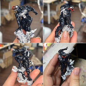 May include: A detailed figurine of the Marvel Comics character Venom. The figure is predominantly black with white accents and a red tongue. It is posed dynamically on a rocky, white base, showcasing intricate detailing.