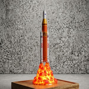 May include: A model of the NASA Space Launch System rocket featuring a central orange core stage, two white side boosters with red NASA lettering, and a white upper stage. The base is surrounded by a glowing, textured orange and yellow flame effect.