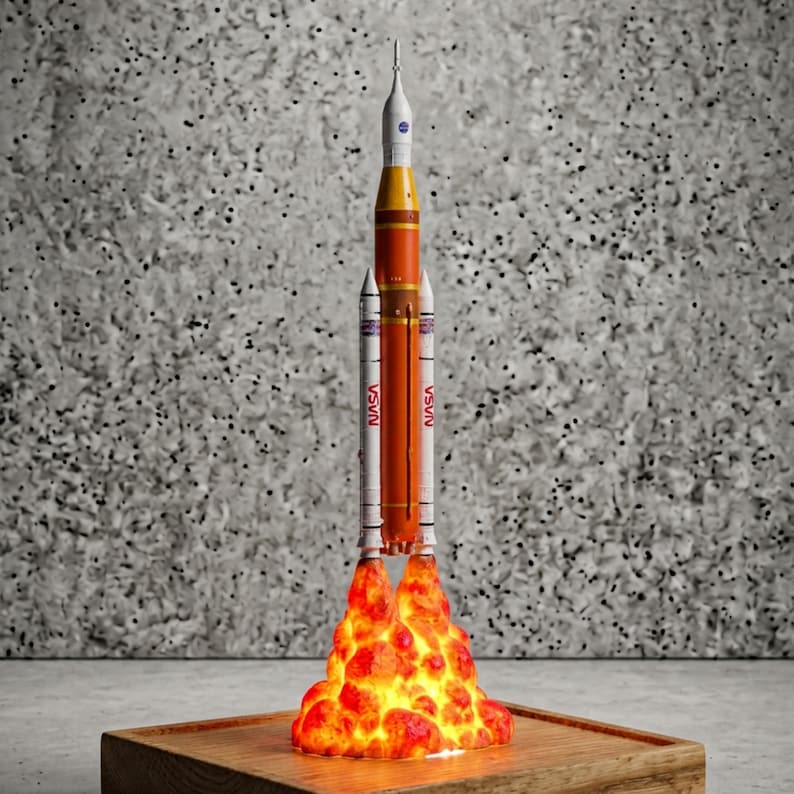 May include: A model of the NASA Space Launch System rocket featuring a central orange core stage, two white side boosters with red NASA lettering, and a white upper stage. The base is surrounded by a glowing, textured orange and yellow flame effect.