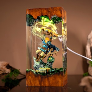 May include: A rectangular resin lamp featuring a wooden base and top, enclosing a miniature scene of a blonde character in a blue tunic holding a bow above a green creature. The internal structure glows with a bright yellow light effect.