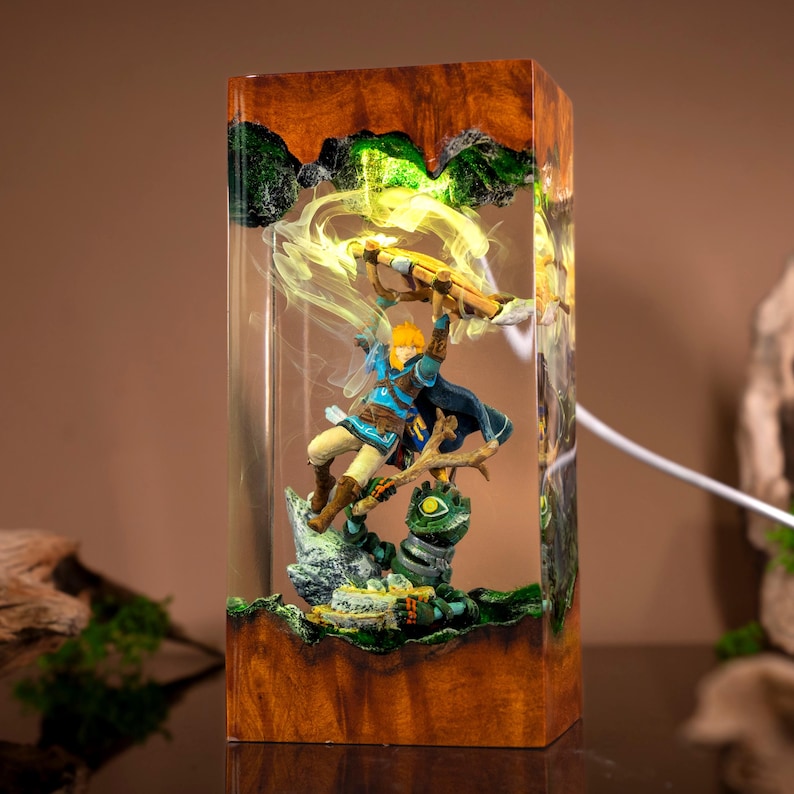 May include: A rectangular resin lamp featuring a wooden base and top, enclosing a miniature scene of a blonde character in a blue tunic holding a bow above a green creature. The internal structure glows with a bright yellow light effect.