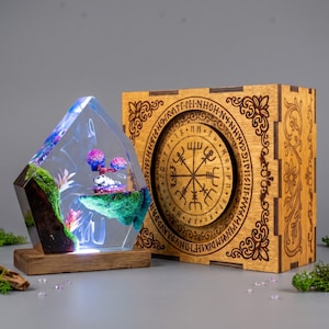 May include: A clear resin lamp shaped like a crystal, containing green moss, pink plants, and a small white figure, set on a wooden base. Beside it is a square wooden box featuring an engraved circular design with a Vegvisir symbol and text.
