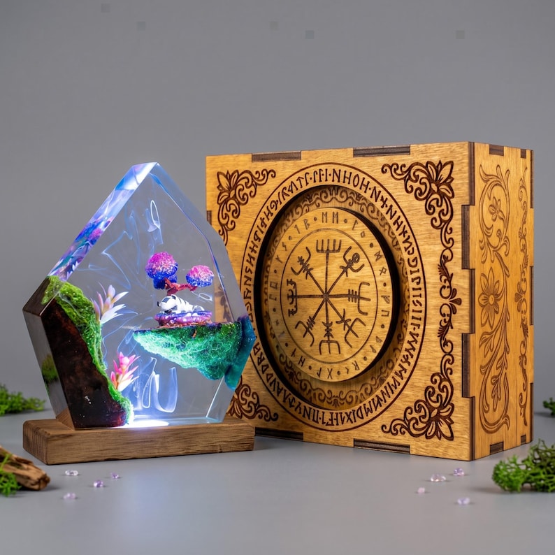 May include: A clear resin lamp shaped like a crystal, containing green moss, pink plants, and a small white figure, set on a wooden base. Beside it is a square wooden box featuring an engraved circular design with a Vegvisir symbol and text.