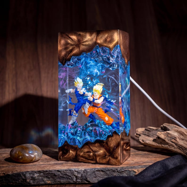Op de afbeelding: Een rechthoekige harslamp met twee anime-figuren in een gevechtshouding, ingesloten in een helder blauw wervelend middenstuk. De boven- en onderkant zijn gemaakt van gestructureerd bronskleurig metaal. Een wit snoer steekt aan de zijkant uit.