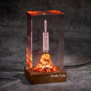 Op de afbeelding: Een helder harsblok met een SpaceX-raketmodel erin, gemonteerd op een houten voet. De raket is wit met zwarte accenten, en de voet heeft een vurige, oranje-gele gloed, die vlammen simuleert. De voet draagt de tekst "Resin Epic".