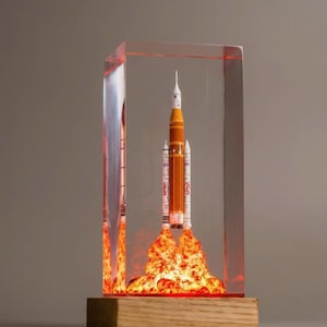May include: A model rocket, orange and white, encased in a clear acrylic block. The rocket is depicted launching with flames at the base. The acrylic block sits on a wooden base.