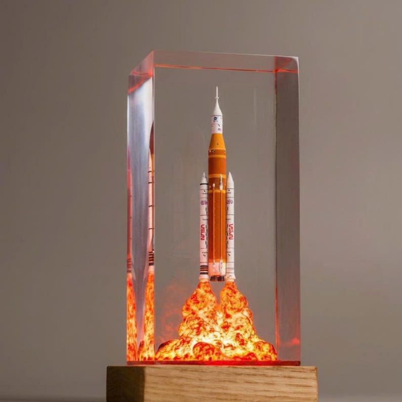 May include: A rectangular clear resin block containing a miniature model of a NASA rocket with an orange central core and two white side boosters. The base of the rocket features a sculpted orange and yellow flame effect, all set on a wooden base.