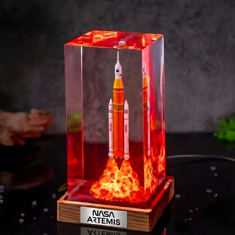 May include: A rectangular resin block containing a miniature NASA Artemis rocket model with a glowing orange light effect at the base, mounted on a wooden stand with a silver plaque displaying the text NASA ARTEMIS in black lettering.