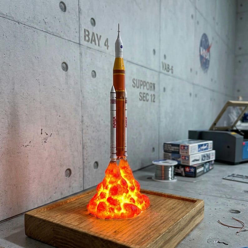 May include: A scale model of the NASA Space Launch System rocket featuring a white and orange cylindrical body with two side boosters. The base of the rocket is illuminated with a bright orange and yellow light effect resembling a fiery exhaust plume.