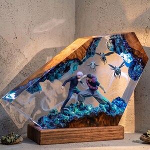 May include: A decorative resin and wood lamp featuring two anime figures in a combat pose. The scene is encased in a translucent blue and clear resin block with embedded blue textures, set upon a rectangular wooden base with internal lighting.