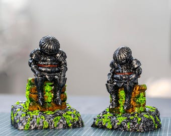 May include: Two miniature figurines of armored knights in dark metallic gray with red waist accents, each sitting on a textured brown stone block base covered in bright green moss, displayed on a green grid cutting mat surface.